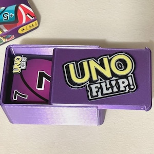 May include: A purple UNO FLIP! card game box with the game's logo in yellow and black. The box is open, revealing a purple card with the number 7. A partial box of UNO cards is in the background.