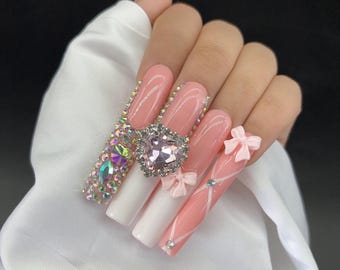 May include: A hand with long, artificial nails. The nails are painted in a combination of pink and white, with decorative elements including rhinestones, bows, and a heart-shaped jewel. The design is intricate and eye-catching.
