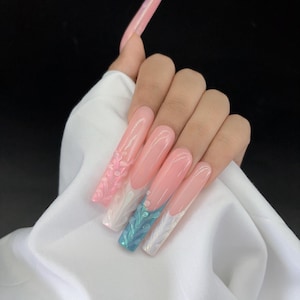 May include: Long, square-shaped artificial nails with a pink base and white tips. One nail features a 3D pink floral design, while another has a blue and white iridescent pattern. The nails are set against a white fabric background.