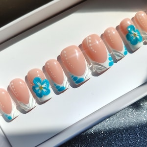 May include: A set of artificial nails with a French manicure design. The nails feature a light pink base, white tips, and blue floral accents. Some nails have white dot patterns. The nails are arranged in a row on a white surface.