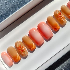 May include: A set of artificial nails in various shades of peach and orange. Some nails feature a floral design with gold accents. The nails are arranged on a white surface, showcasing a gradient effect.
