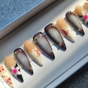 Puede incluir: Un conjunto de uñas postizas largas en forma de almendra. Las uñas presentan una variedad de diseños, incluyendo un degradado de marrón y gris, beige liso y beige con lunares rojos. Algunas uñas tienen adornos dorados y diseños florales.