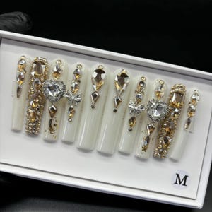 May include: A set of long, white artificial nails, each adorned with an array of sparkling rhinestones in gold and clear hues. Some nails feature heart and butterfly accents, creating a glamorous and eye-catching design. The nails are arranged in a white box with an "M" label.