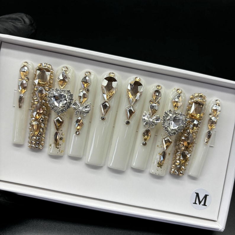May include: A set of long, white artificial nails, each adorned with an array of sparkling rhinestones in gold and clear hues. Some nails feature heart and butterfly accents, creating a glamorous and eye-catching design. The nails are arranged in a white box with an "M" label.