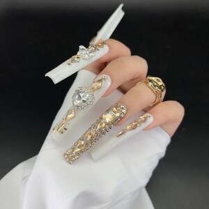 May include: A hand with long, white, embellished acrylic nails. The nails feature various gold and silver rhinestones, including heart and diamond shapes. A gold ring is also visible.