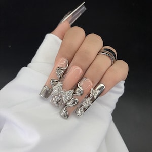 May include: A hand with long, sculpted artificial nails. The nails are a nude color with silver embellishments, including wavy designs, bows, and butterflies. Some nails have clear extensions with silver accents. A silver ring is on the ring finger.