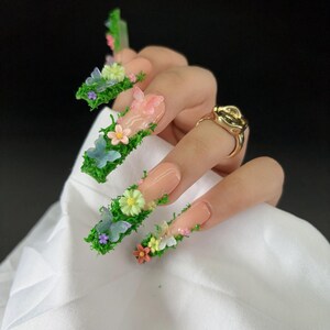 May include: Long, square-shaped artificial nails with a nude base, adorned with green moss, miniature flowers, and butterfly accents. A gold ring is visible on the ring finger. The nails are a unique and artistic fashion statement.