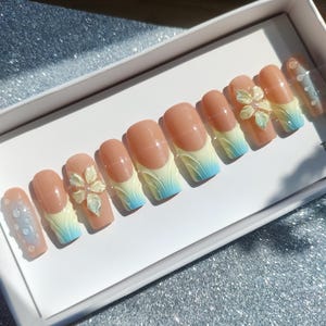 May include: A set of artificial nails in a white box. The nails are a light peach color with a French tip design that fades from yellow to blue. Some nails feature a 3D flower design.