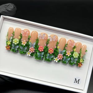 May include: A set of artificial nails in a white box. The nails are a nude color, decorated with green moss, colorful flowers, and butterfly accents. The box has the letter "M" in the bottom right corner.