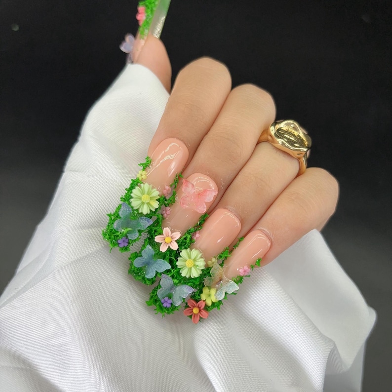 May include: Long, square-shaped artificial nails with a nude base, adorned with green moss, colorful flowers, and butterflies. A gold ring is visible on the ring finger. The nails are a unique and artistic fashion statement.
