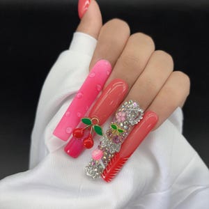 May include: A hand with long, artificial nails. The nails are painted in shades of pink and red, with decorative elements including cherries, rhinestones, and floral designs. One nail has a dotted pattern, and another has a red French tip.