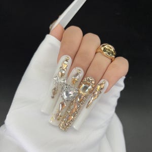 Può includere: Lunghe unghie acriliche bianche decorate con strass dorati e trasparenti, tra cui un design a forma di cuore e un fiocco. È visibile anche un anello d'oro. Le unghie sono su uno sfondo di tessuto bianco.