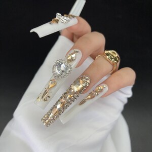May include: Long, white acrylic nails adorned with gold and clear rhinestones. One nail features a heart-shaped rhinestone, while others have a mix of gold accents and various rhinestone shapes. A gold ring is also visible.