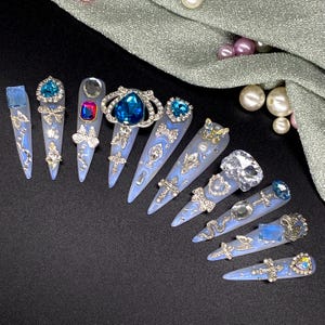 Könnte beinhalten: Eine Sammlung hellblauer, stilettoförmiger Kunstnägel, die jeweils mit aufwendigen silbernen Verzierungen und funkelnden Edelsteinen verziert sind. Designs umfassen Schleifen, Kreuze und verschiedene Juwelenakzente, die eine glamouröse Ästhetik schaffen.