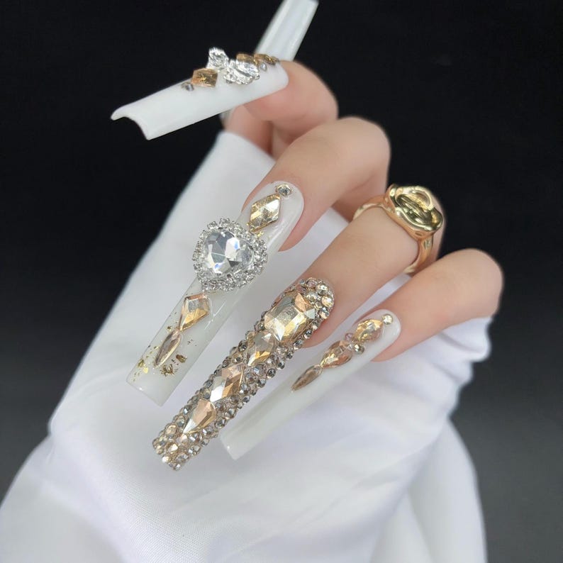 May include: White, long, square-shaped artificial nails adorned with gold and clear rhinestones. One nail features a heart-shaped rhinestone. A gold ring is visible on a finger. The nails are a fashion statement.