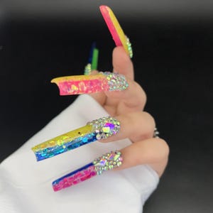May include: Long, colorful acrylic nails with a gradient design. The nails transition from yellow to pink to blue, with glitter and rhinestones. The nails are square-shaped and have a glossy finish.