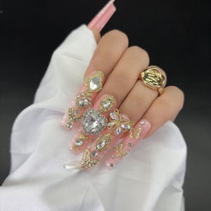 May include: Long, pink acrylic nails adorned with gold and rhinestone embellishments. The designs include butterflies, hearts, and geometric shapes. A gold ring with a textured design is also visible.
