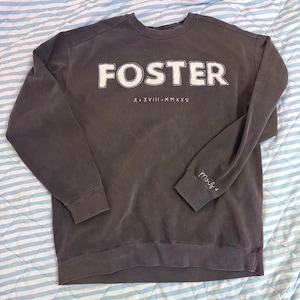 May include: A charcoal gray crewneck sweatshirt featuring the word FOSTER in large white stitched lettering across the chest. Below the name, the Roman numerals X. XVIII. MMXXV are embroidered in white. The left sleeve cuff displays the word mrs.