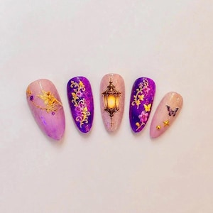 May include: Five almond-shaped press-on nails featuring a purple and light pink gradient base with glitter. Designs include a gold fairy, gold floral vines, a glowing lantern, purple flowers, a small butterfly, and tiny gold metallic studs.
