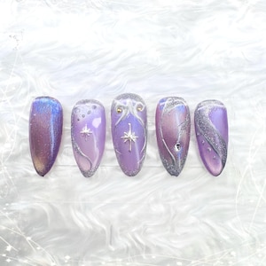 May include: Five almond-shaped artificial nails with a purple and silver design. Each nail features a different design, including glitter, stars, and swirls. The nails have a glossy finish and are set against a white, fluffy background.