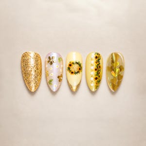 May include: Five almond-shaped press-on nails featuring diverse designs: one solid gold glitter, one iridescent with white flowers, one pale yellow with a sunflower wreath, one yellow with pearls and sunflowers, and one with gold heart accents.