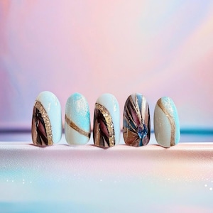 May include: Five press-on nails featuring a light blue base with decorative accents. Designs include gold glitter lines, dark purple and brown abstract strokes, and a central blue gemstone embellishment on one nail, creating a cohesive set.