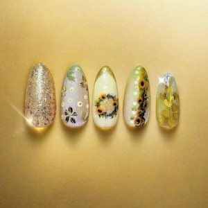 May include: Five almond-shaped artificial nails with various designs. The first nail is covered in gold glitter. The second nail has a floral design with butterflies. The third nail features a sunflower wreath. The fourth nail has sunflowers and gold dots. The last nail has gold heart-shaped glitter.