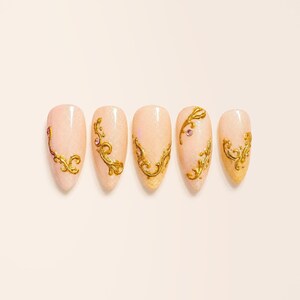 May include: Five almond-shaped press-on nails with a pale peach base colour. Each nail features raised gold-coloured filigree scrollwork designs and small embedded pink rhinestones, arranged in a row against a light background.