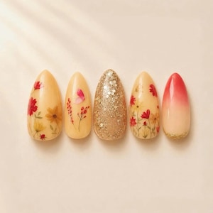 May include: Five almond-shaped press-on nails arranged in a row. The first, second, and fourth nails feature a nude base with pressed red and yellow flowers. The third nail is covered in gold glitter, and the fifth has a red-to-nude gradient.