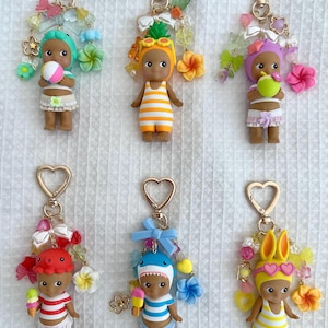 Puede incluir: Seis llaveros con pequeñas figuras vestidas con varios disfraces, incluyendo una piña, un tiburón y un pulpo. Cada figura está unida a un cierre dorado en forma de corazón y decorada con cuentas coloridas, lazos y dijes en forma de flor.