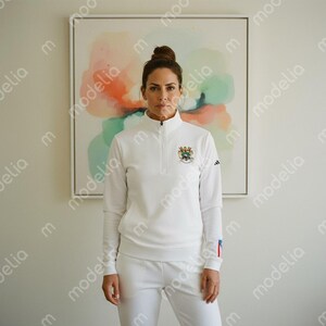 May include: White athletic tracksuit featuring a long-sleeve top with a quarter-zip collar and a small emblem on the chest. The matching pants complete the set. The Adidas logo is visible on the sleeve. The outfit is worn by a person.