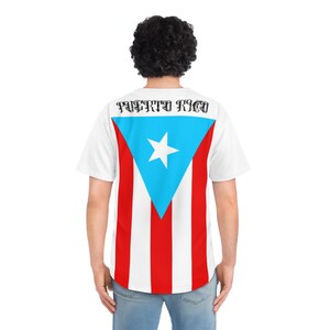 May include: White baseball jersey featuring the Puerto Rican flag design on the back. The flag includes a blue triangle with a white star, and red and white stripes. The word "PUERTO RICO" is printed across the top of the jersey.
