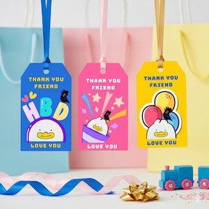 以下が含まれることがあります： 青、ピンク、黄色の背景に、3つのカラフルなギフトタグ。各タグにはリボンが付いており、「THANK YOU FRIEND」と「LOVE YOU」の文字が書かれています。青いタグには「HBD」と漫画のアヒルが描かれています。ピンクのタグには星とアヒルが描かれています。黄色のタグには風船とアヒルが描かれています。