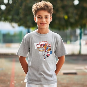 Puede incluir: Una camiseta gris con un gráfico de baloncesto y las palabras "HOOP LOOPS". El diseño muestra un personaje de dibujos animados encestando una pelota de baloncesto. La camiseta es de manga corta.