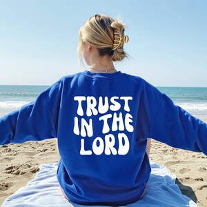 Könnte beinhalten: Ein königsblaues Sweatshirt mit dem weißen Schriftzug "TRUST IN THE LORD" auf dem Rücken. Das Sweatshirt wird von einer Person am Strand getragen, mit Meer und Himmel im Hintergrund.