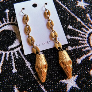 Puede incluir: Pendientes de cabeza de serpiente dorados con un diseño de cadena. Los pendientes están en una tarjeta blanca con el texto "Dani Awesome" impreso en ella.