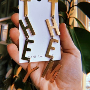 Puede incluir: Pendientes colgantes de tono dorado con las letras "THEM" colgando de cada pendiente. Los pendientes están en una tarjeta blanca con el texto "Dani Awesome" impreso en ella.