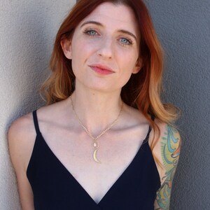 May include: A woman with red hair wearing a black sleeveless top and a gold necklace with a crescent moon pendant. She is standing in front of a white wall.