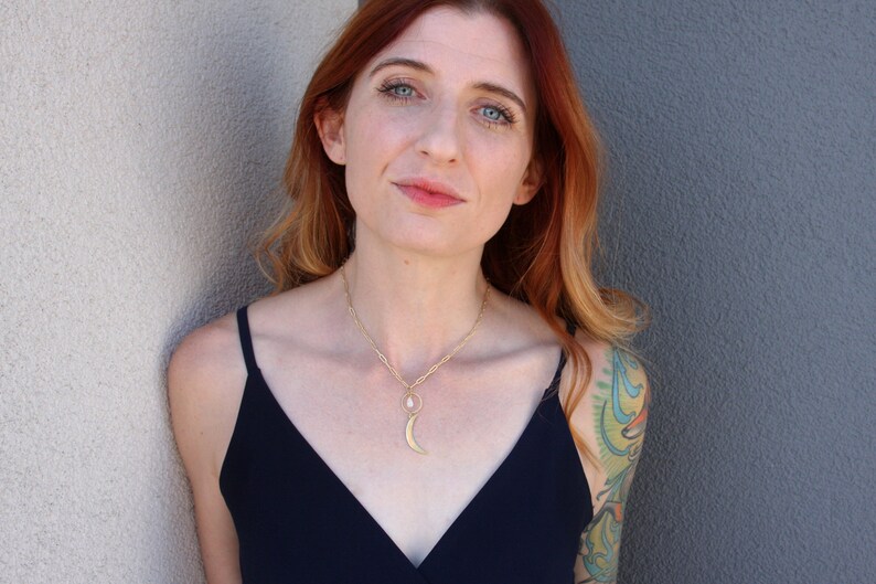 May include: A woman with red hair wearing a black sleeveless top and a gold necklace with a crescent moon pendant. She is standing in front of a white wall.