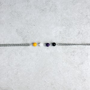 Op de afbeelding: Een zilveren ketting met vier kralen: geel, wit, paars en zwart. De kralen zijn gelijkmatig verdeeld over de ketting.