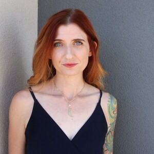 May include: A woman with long, wavy red hair wearing a black sleeveless top and a gold necklace with a crescent moon pendant. She has a colorful tattoo on her right arm.