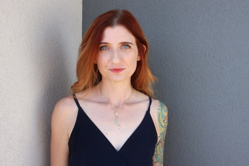 May include: A woman with long, wavy red hair wearing a black sleeveless top and a gold necklace with a crescent moon pendant. She has a colorful tattoo on her right arm.