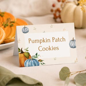May include: A rectangular folded paper sign with a cream background featuring watercolor illustrations of orange and blue pumpkins and green leaves. The text "Pumpkin Patch Cookies" is printed in the center in a brown serif font.