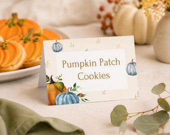 May include: A rectangular folded paper sign with a cream background featuring watercolor illustrations of orange and blue pumpkins and green leaves. The text "Pumpkin Patch Cookies" is printed in the center in a brown serif font.