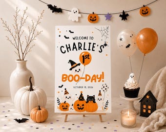 May include: A rectangular white sign on a wooden easel featuring the text WELCOME TO CHARLIE'S 1st BOO-DAY! OCTOBER 18, 2026. The design includes illustrations of ghosts, a black cat, orange pumpkins, bats, and a spider web in orange and black.