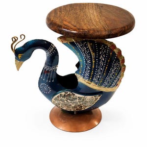 Puede incluir: Una mesa auxiliar decorativa con forma de pavo real. La mesa tiene un cuerpo azul oscuro con detalles pintados en blanco y dorado, una base de color cobre y una tapa redonda de madera. La cabeza del pavo real está erguida, con pico dorado y adornos.