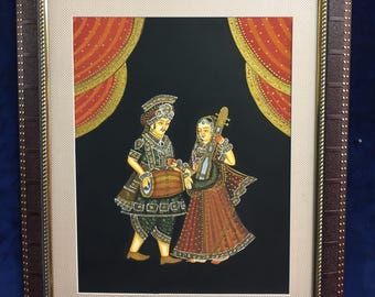 May include: Framed artwork depicting two figures in traditional attire, one playing a drum and the other a stringed instrument. The figures stand against a black backdrop with red and gold curtains. The frame is brown with a textured border.