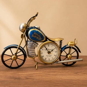 May include: A blue and gold motorcycle-shaped clock. The clock face is round and gold-rimmed, with the words "Vintage Clockworks" on it. The words "Royal Enfield" are on the fuel tank. The wheels are black.