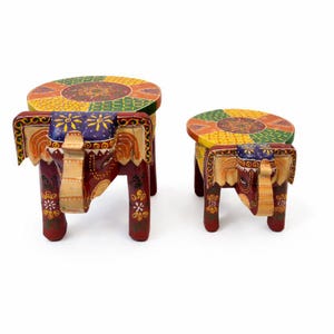 Puede incluir: Dos taburetes decorativos de madera con forma de elefante. Los taburetes están pintados con colores vibrantes, incluyendo rojo, amarillo, verde y morado. Las patas con forma de elefante y las tapas redondas presentan intrincados diseños florales. El taburete más grande mide unos 30 cm de altura.
