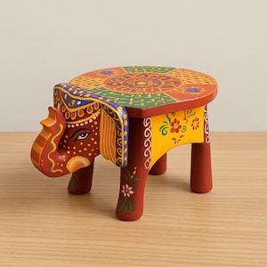Puede incluir: Un taburete de elefante de madera pintado a mano. El taburete presenta una parte superior redonda con un diseño floral y geométrico colorido. La cabeza y las patas del elefante están pintadas en tonos de rojo, amarillo y marrón, con detalles intrincados.